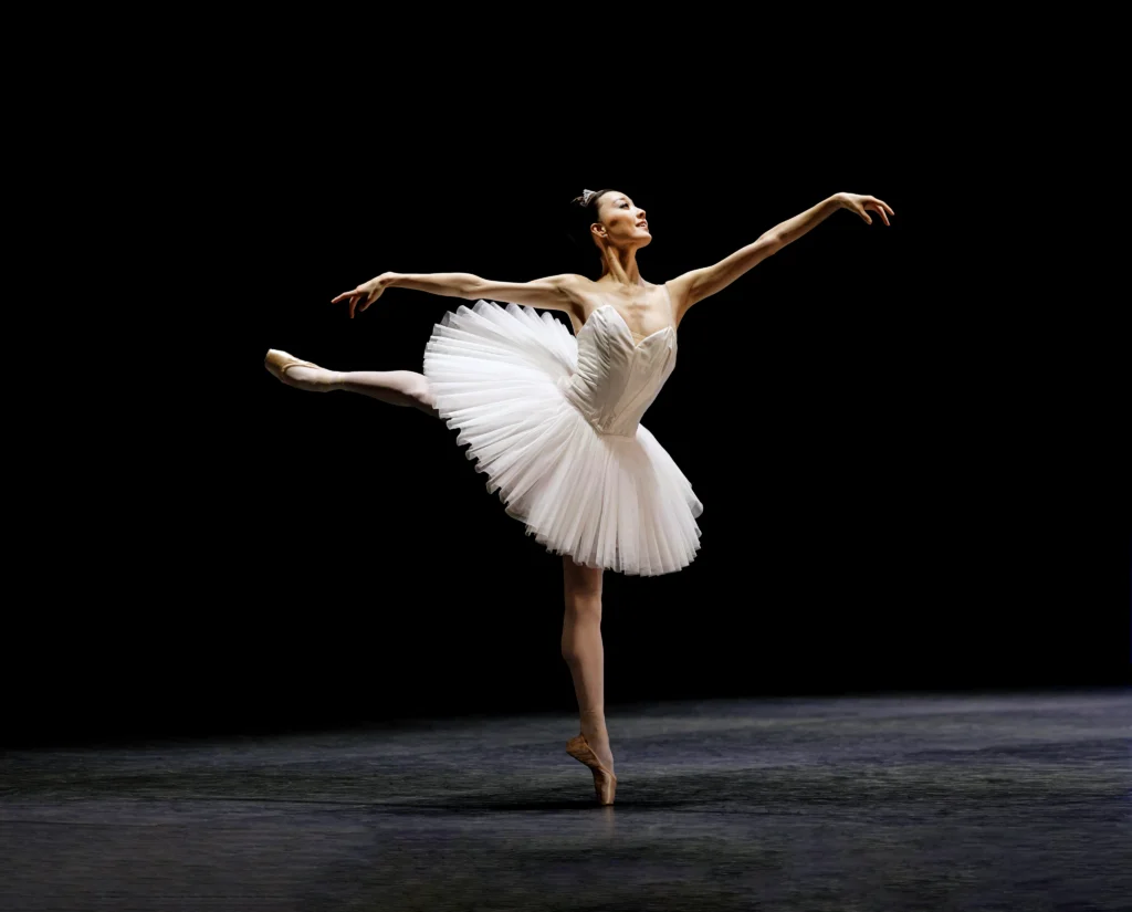 Hohyun Kang piques to first arabesque on a shadowy stage, a subtle smile on her face. She wears a simple white tutu, pink tights, and pointe shoes.