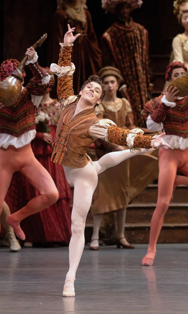 Jake Roxander piques to croisé attitude back, palms open in high fifth and second. He smiles easily, chin raised. He wears an orange-brown tunic with white poofs along the sleeves, white tights, and ballet slippers. Similarly costumed dancers with prop mandolins and watching villagers are visible upstage.