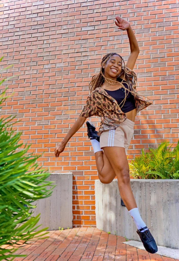 Kaitlyn Sardin smiles sunnily as she flies through the air. Her legs are tight together, one heel tucked up behind her, the opposite arm tossed overhead. She wears a brown, geometrically patterned blouse open over a black sports bra and beige athletic shorts. Her blonde and brown braids fly around her.