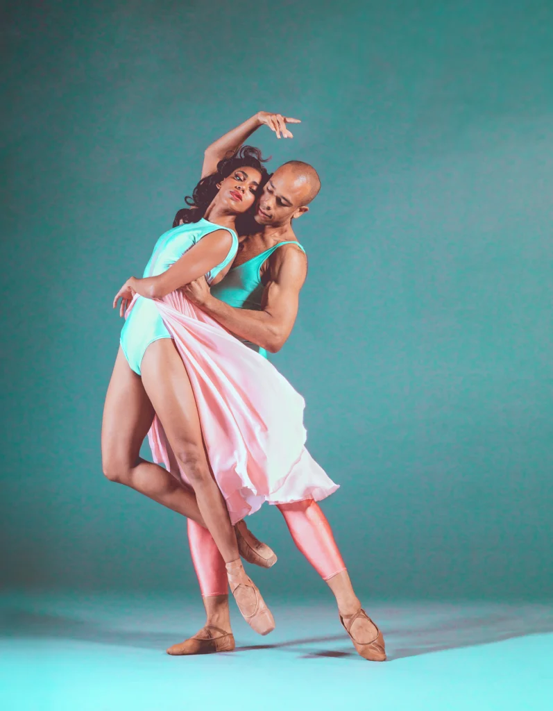 Kamala Saara is lifted a few inches off the floor by the waist, legs in coupé back. One arm twists across her waist, the other in high fifth. Her dark hair curls around her face as she turns her head toward her partner. She wears a teal leotard and a flowing pastel, pink skirt, no tights, and pointe shoes painted to match her complexion.