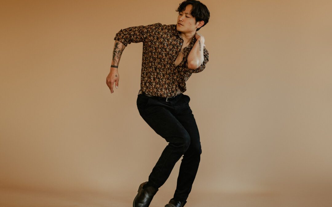 a male dancer wearing black pants and a patterned shirt posing against a tan backdrop