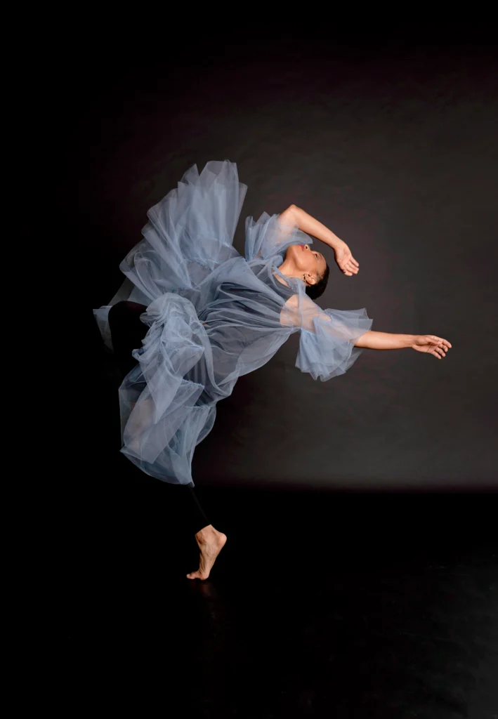 An African American woman on a black background dances wearing a blue flowing dress. She arches backward with one leg bent, one arm extended and the other arm bent above her head. Her eyes are closed.