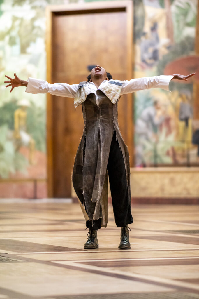 a dancer wearing a long brown costume reaching out and looking up