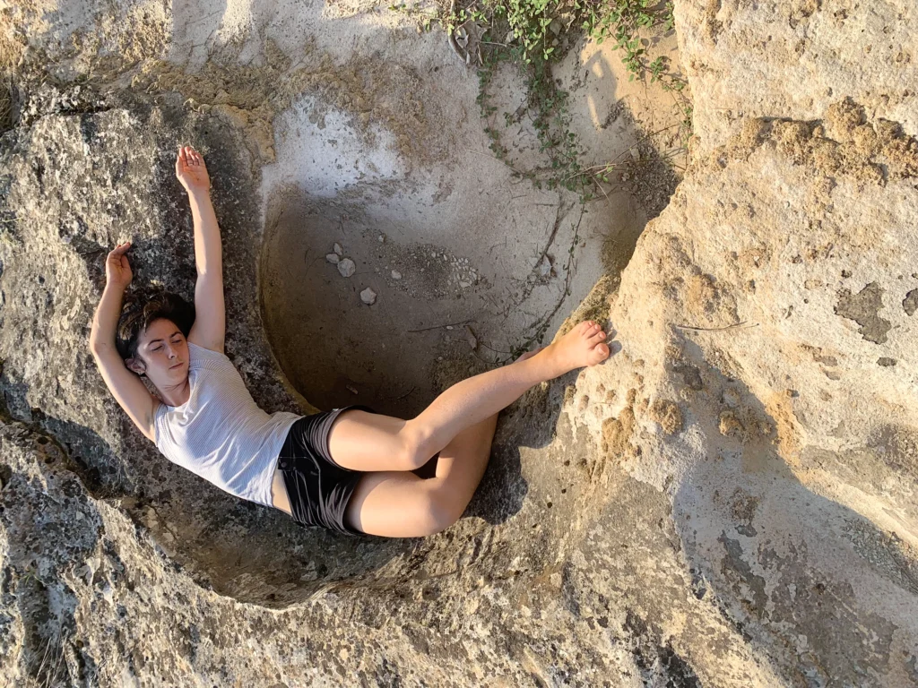 Lucy Fandel lies on her back, arching to match the curving of the rock around and beneath her. Her eyes are closed, arms draping overhead, while her bare feet press against the edge of the rock. She wears a simple white t-shirt and black shorts.