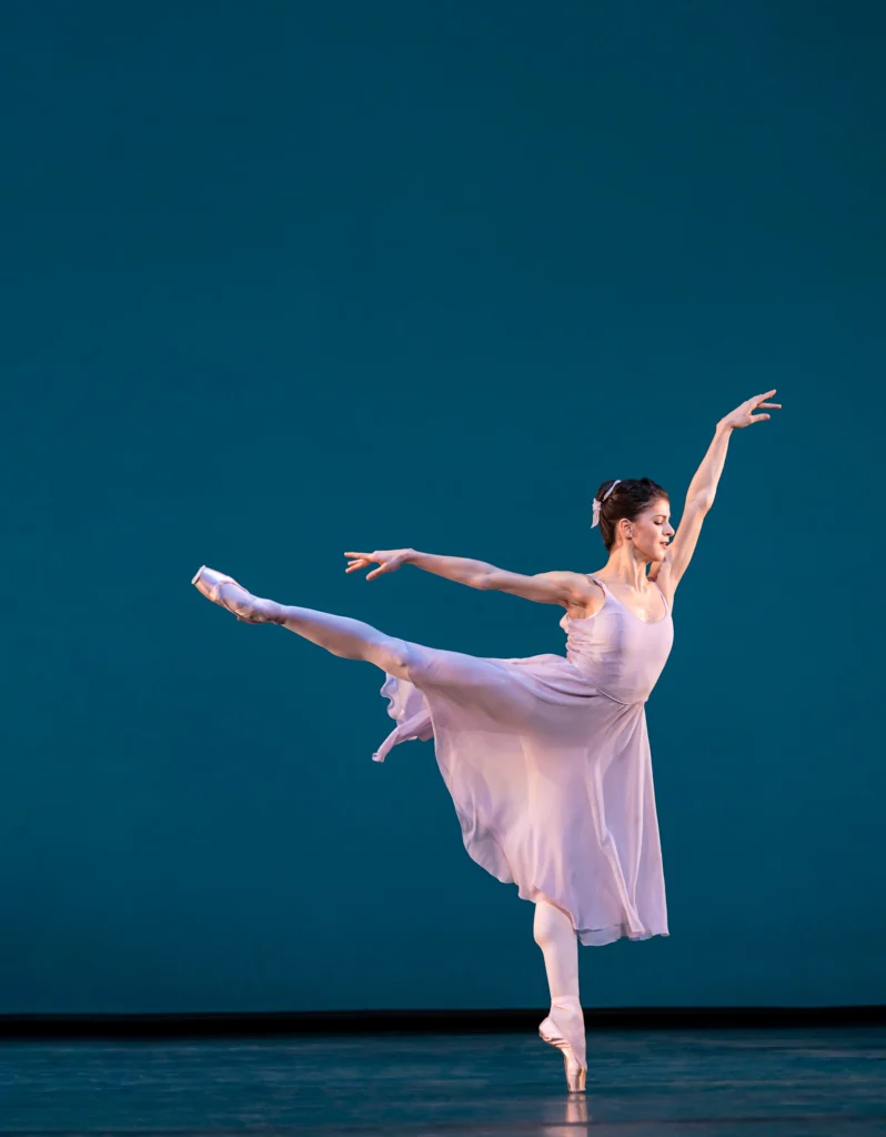Mayara Magri balances in first arabesque on an otherwise empty stage against a deep blue backdrop. She wears a pale pink dress that falls just past her knee, pink tights, and pointe shoes.