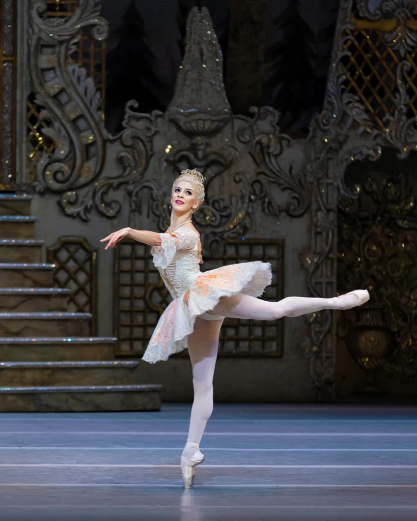 Mayara Magri in performance. She poses in back attitude on pointe, at a clean 90 degrees. Her arms are at shoulder height, sweeping through second arabesque. She wears a classical tutu with half sleeves that drape at the elbows, pink tights and pointe shoes, and a white wig topped by a tiara.
