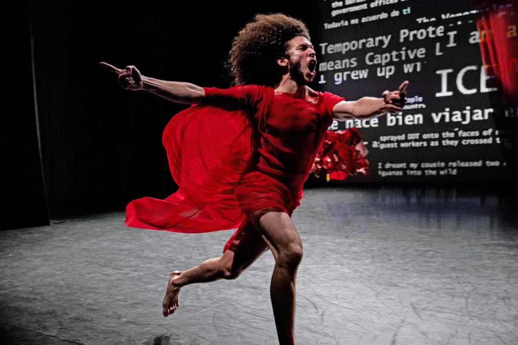 Miguel Alejandro Castillo runs, mouth wide open seeming to yell. His arms are outstretched, pointer fingers aiming ahead and to the side. His puffy hair flies behind him, as does the draping fabric of his red costume. Words in white font on a black backdrop are projected on the back wall.