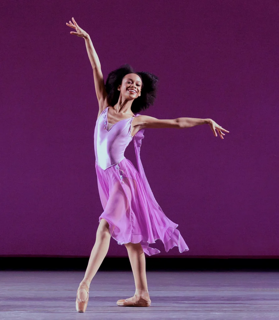 Olivia Bell poses in tendu croisé devant. One arm is extended side, the other by her head. She gives a radiant smile, natural hair framing her face. She wears a purple, flowing dress over tights and pointe shoes.