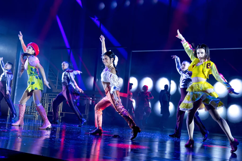 Pauline Casiño, with braided hair and wearing a white crop top and pink pants, poses with her right arm pointing diagonally upwards onstage in the Broadway musical Once Upon a One More Time.