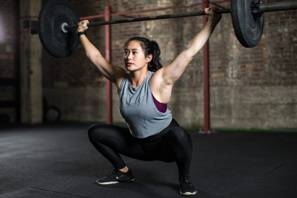 a woman in a grey tank top squatting while holding a barbell over her head