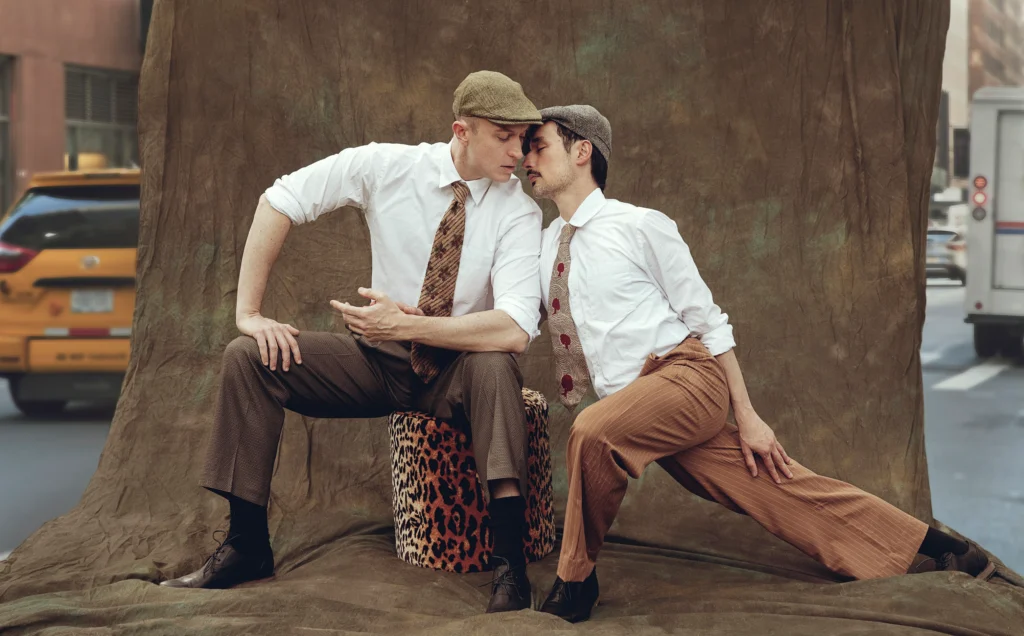Two male dancers in ties, dress shirts with the sleeves rolled, and flat caps pose together against a photo backdrop set up outdoors. One sits on a block, touching his forehead to the other dancer's as he lunges alongside.