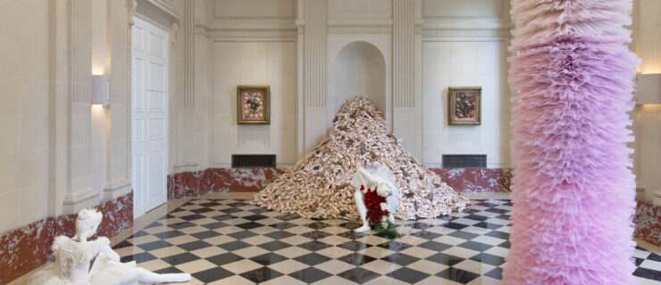 A pile of pointe shoes and a tower of pink tutus frame a formal, marble-walled and marble-floored room. Two white plaster statues of ballet dancers anchor the scene. One kneels, bowing, in front of the pile of pointe shoes; the other sits off to the left of the frame, adjusting her shoe.