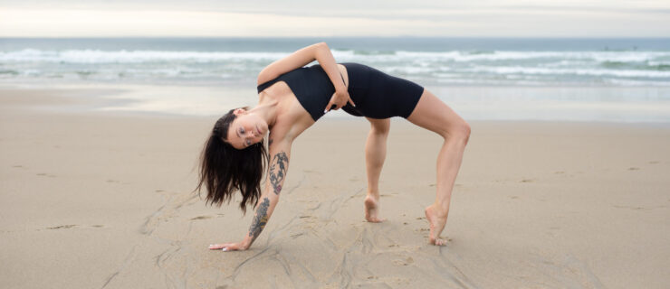The author, a white woman with dark hair and tattoos on her right arm, dancing on the beach, suspended in a forced arch bridge shape with her left arm wrapped around her torso.