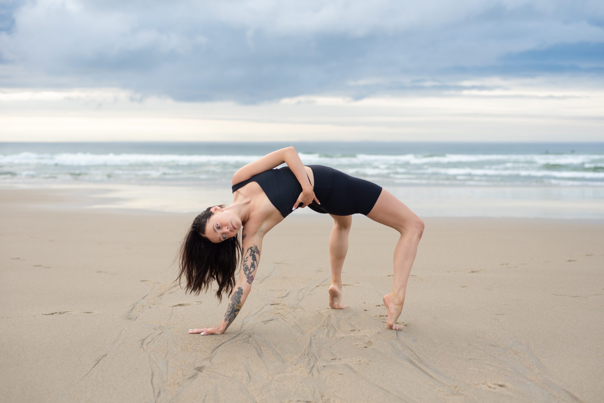 The author, a white woman with dark hair and tattoos on her right arm, dancing on the beach, suspended in a forced arch bridge shape with her left arm wrapped around her torso.