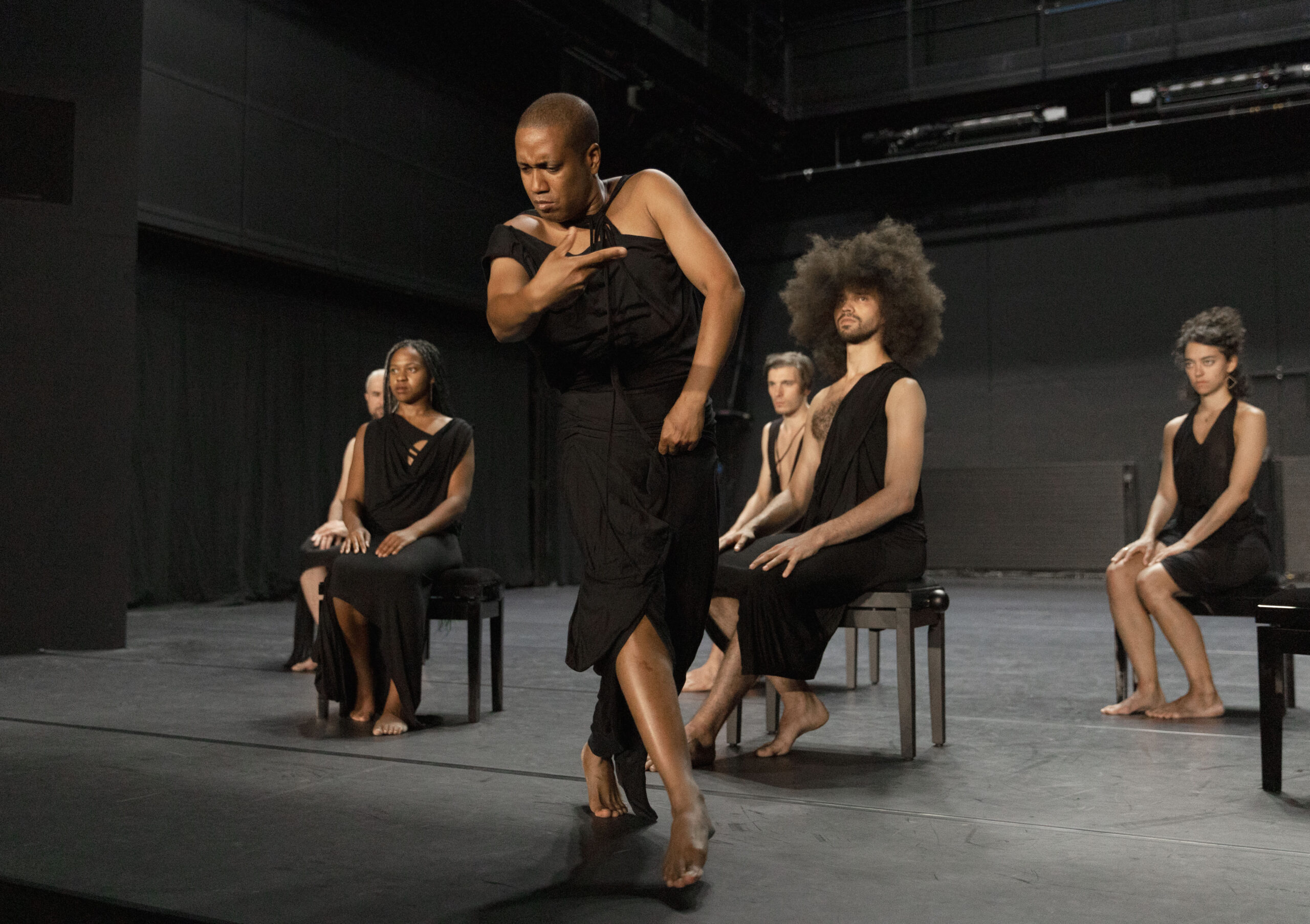 a dancer wearing a black dress performing downstage; a group of other dancers sitting in chairs behind