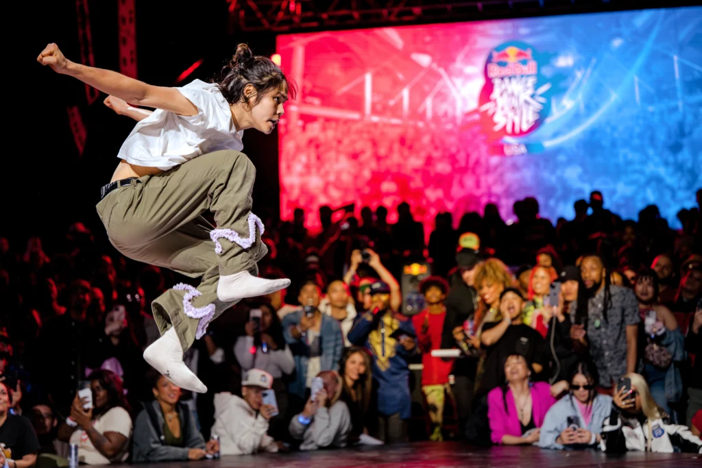 Sean Lew, a dancer in a white t-shirt, olive pants with pink trimming, and off-white socks, competes at the Red Bull Dance Your Style National Finals in Chicago on May 20, 2023. He is jumping in the air, with his fists stretched behind him and his knees pulled to his chest.