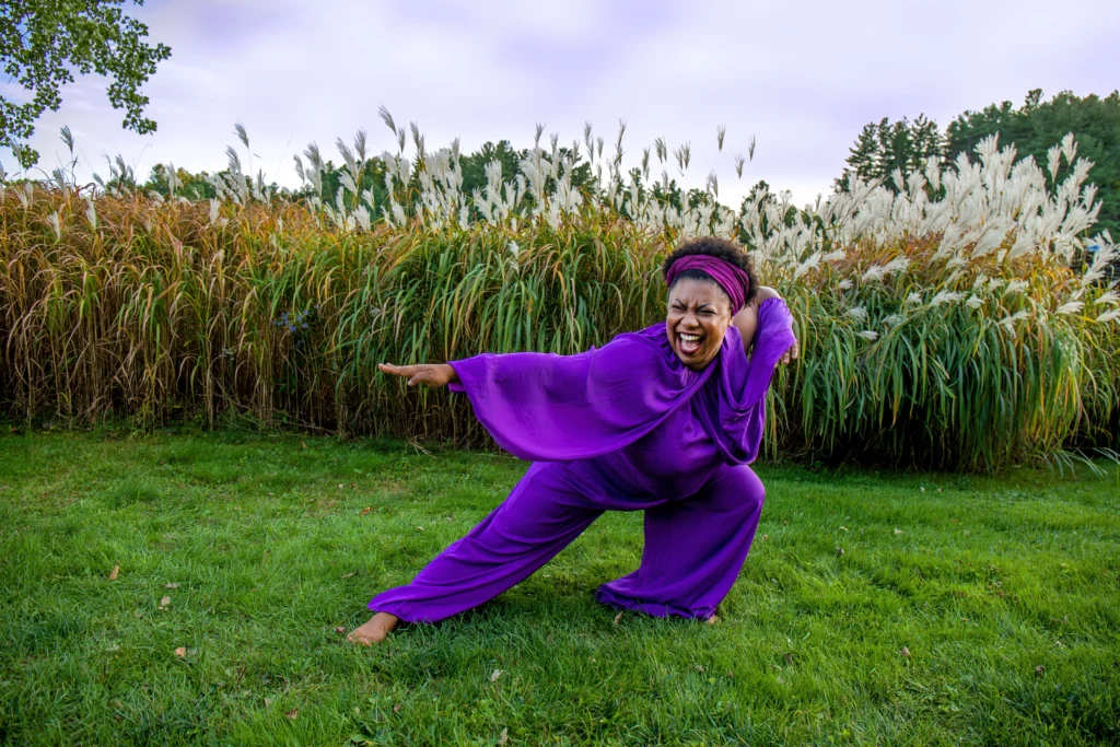 Sydnie Mosley, a Black woman wearing a flowy purple jumpsuit lunges back with her arms out. Her short black afro is held back by a purple scarf, her face shows a clear expression of joy. She is standing barefoot in front of the natural background of Ashfield, Massachusetts. 
