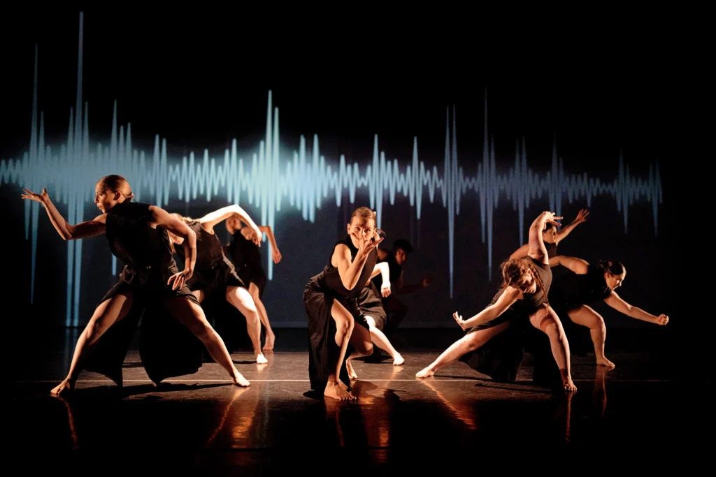 Six dancers lunge out of a square of light, each raising a splayed hand as though catching something from the air. Visual representation of a soundwave is projected on the back wall. They are costumed in black tank tops and wide legged pants slit up to the mid-thigh.