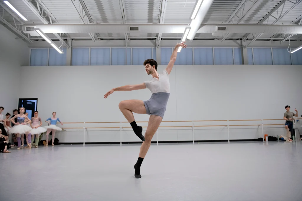 Yuval Cohen in retiré passé, arms in an elegant L as he tips slightly off balance. He is in the center of a large rehearsal studio, wearing a white and blue biketard and black ballet slippers.