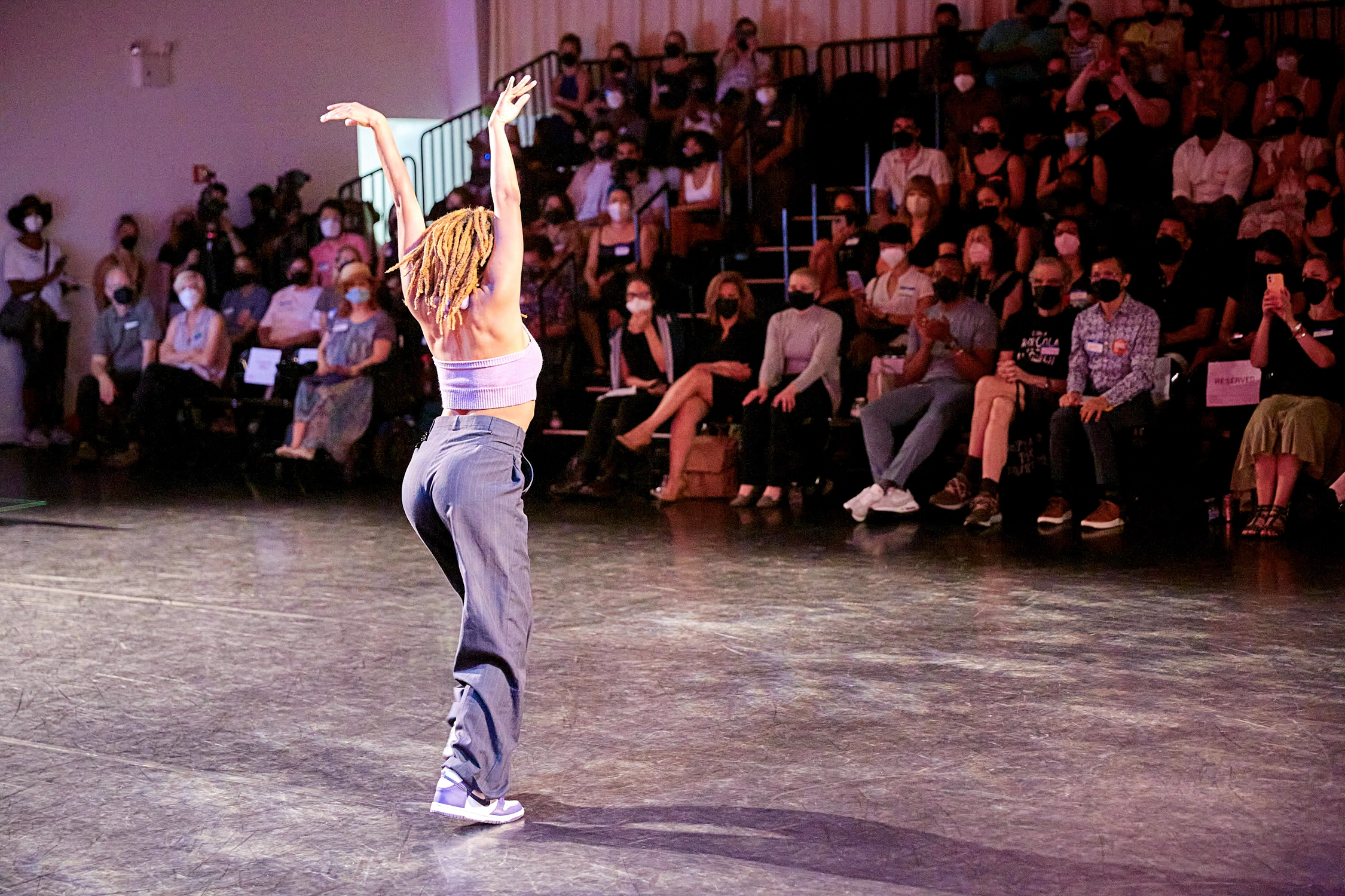 A dancer poses with their back to the camera, arms in a V overhead. A masked audience seated on risers watches.