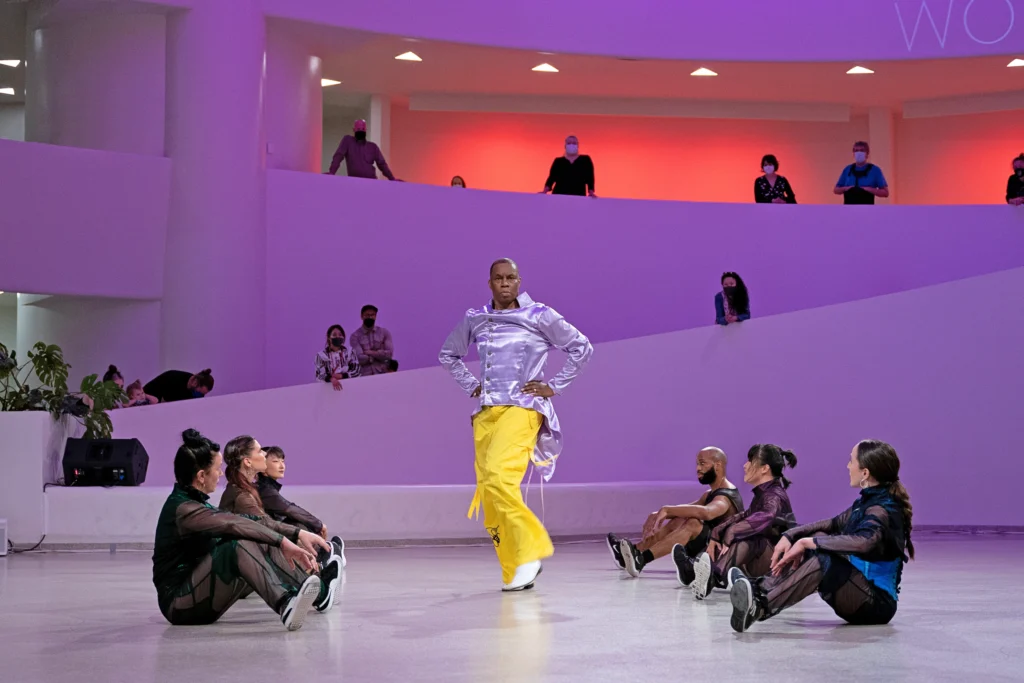 Archie Burnett stands with his hands on his hips and one foot popped, wearing bright yellow pants and a shiny top. Six dancers in black sit in two lines of three on either side of him, looking to him as they form an aisle. The ramp that curves around the Guggenheim Rotunda is dotted with audience members every five or six feet.