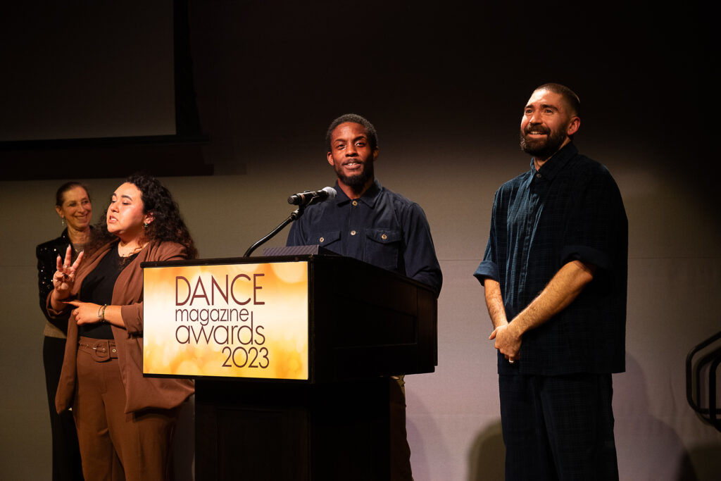 Amadi "Baye" Washington, a young Black man, speaks at a podium with a microphone and a sign that reads "Dance Magazine Awards 2023." Sam "Asa" Pratt, a young white man with a bushy beard, stands beside him smiling, hands clasped in front of him. Joan Finkelstein looks on smiling in the near background, as an ASL interpreter signs in the foreground.