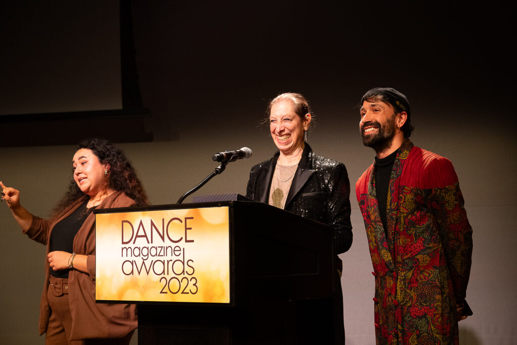 Joan Finkelstein smiles broadly as she stands at a podium with a sign that reads "Dance Magazine Awards 2023." Beside her is a smiling Omar Román De Jesús, a young Latino man wearing a red robe with a floral pattern over a black sweater.