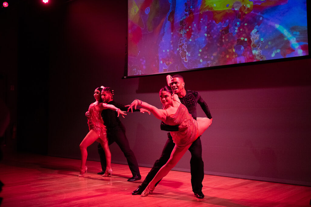 Two couples performing a Latin ballroom routine pose on a stage awash in red light. 