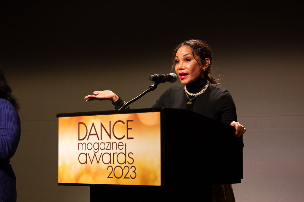 Daphne Rubin-Vega, a Latina woman, gestures expressively as she speaks into a microphone at a podium with a sign that reads "Dance Magazine Awards 2023."