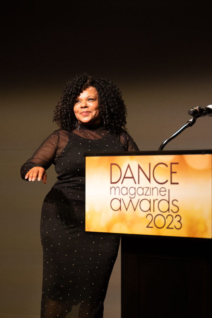 Maria Torres, a curvy Latina woman, gives a close-lipped smile as she turns in place to show off a sparkly, form-fitting black dress. She is beside a podium with a sign that reads "Dance Magazine Awards 2023."