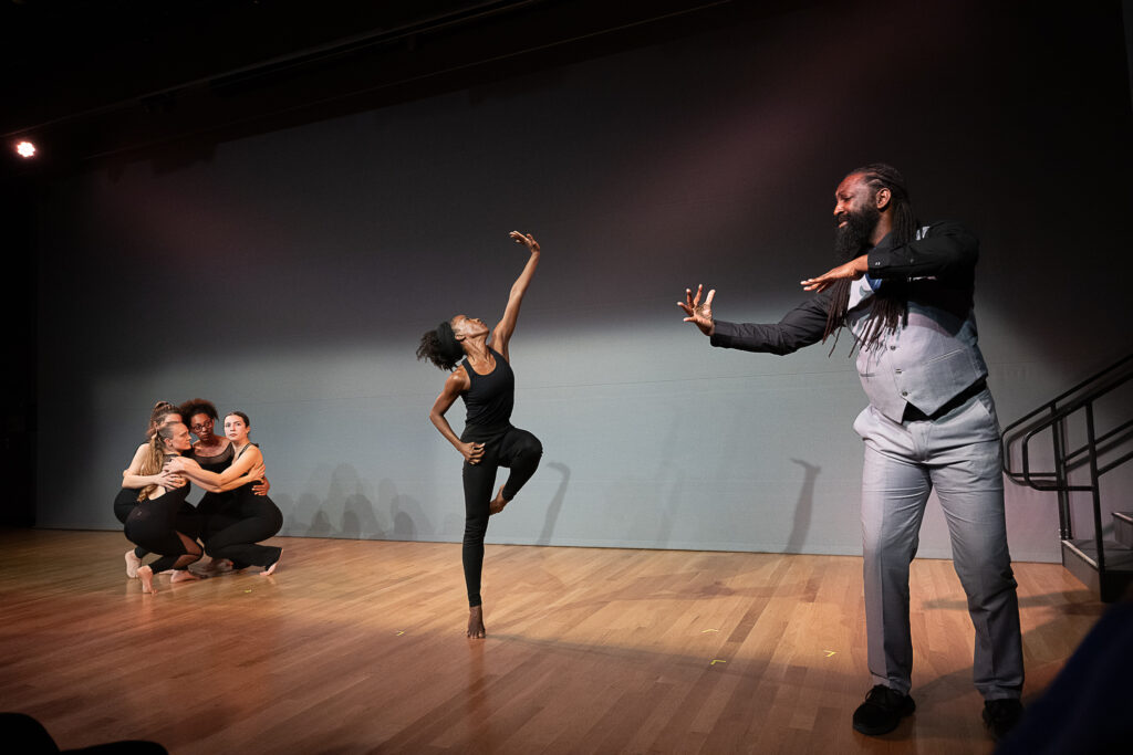 Antoine Hunter, a Black and Indigenous person with long dreads and beard, closes his eyes as he signs the music downstage. A Black dancer at center stage balances in parallel passé, one hand pressing the sky away as she looks up to it. A cluster of four dancers crouch together upstage, arms around each other's backs as they look to the dancer at center.