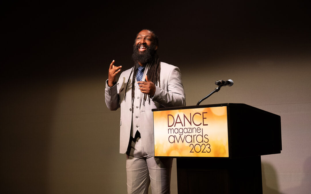 Antoine Hunter Purplefirecrow, a Black and Indigenous person with long dreads and a bushy beard. He wears a sharp, pale grey suit. He gives an infectious smile as he signs, standing beside a podium emblazoned with the logo for the 2023 Dance Magazine Awards.