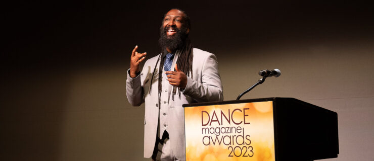 Antoine Hunter Purplefirecrow, a Black and Indigenous person with long dreads and a bushy beard. He wears a sharp, pale grey suit. He gives an infectious smile as he signs, standing beside a podium emblazoned with the logo for the 2023 Dance Magazine Awards.