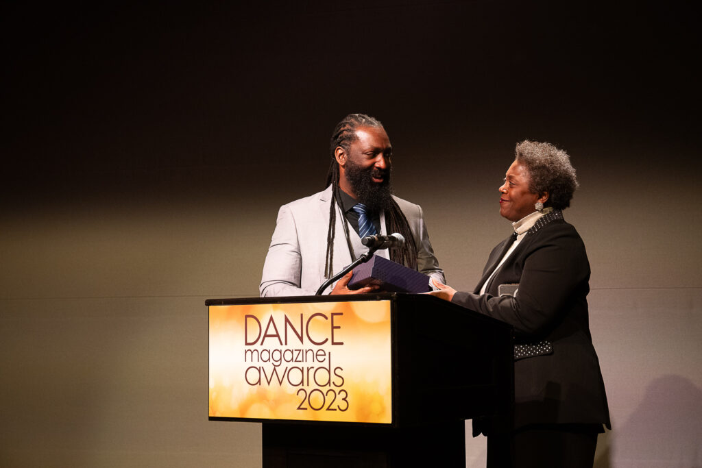 A teary-eyed Antoine Hunter Purplefirecrow, a Black and Indigenous person with long dreads and beard, accepts a box holding a Dance Magazine Award from an emotional Denise Saunders Thompson, an older Black woman. The two share a sense of warmth as they maintain eye contact.
