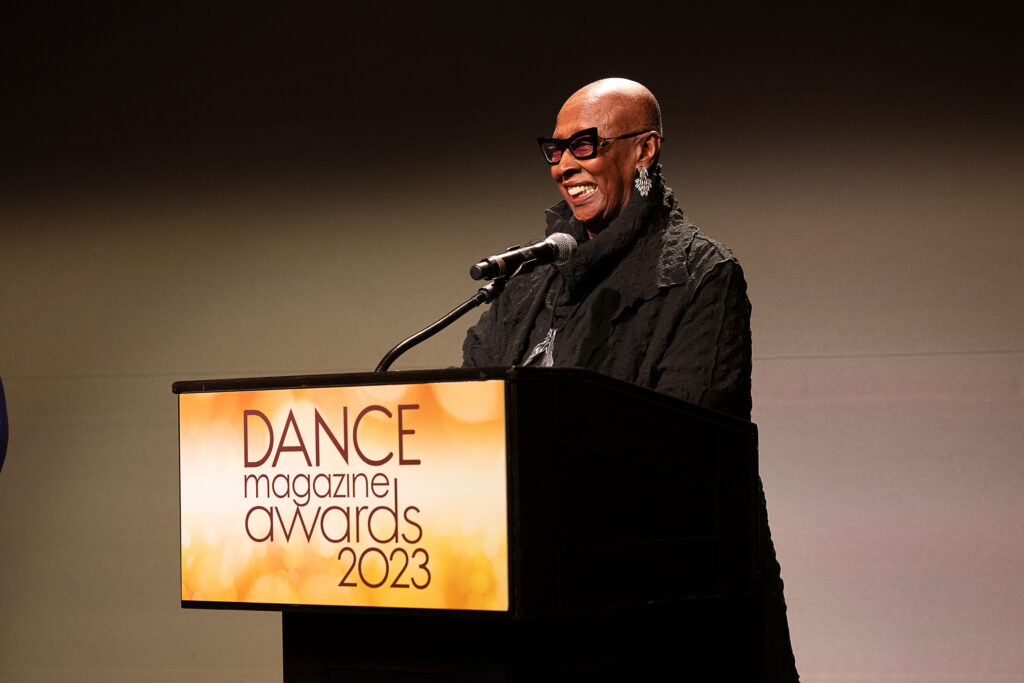 Judith Jamison, an older Black woman with a shaved head and angular tinted glasses, smiles radiantly as she speaks into a microphone at a podium with a sign that reads "Dance Magazine Awards 2023."