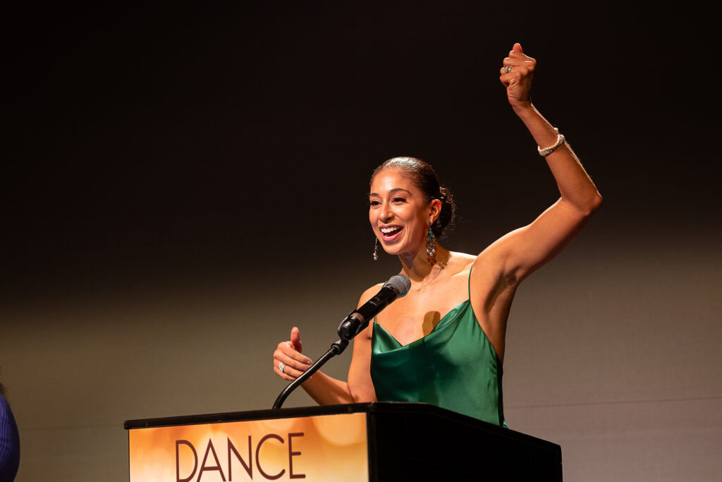 Alicia Graf Mack smiles and laughs as she stands behind a podium. She raises one arm as though holding an imaginary umbrella. She wears an elegant green gown and dangly earrings.