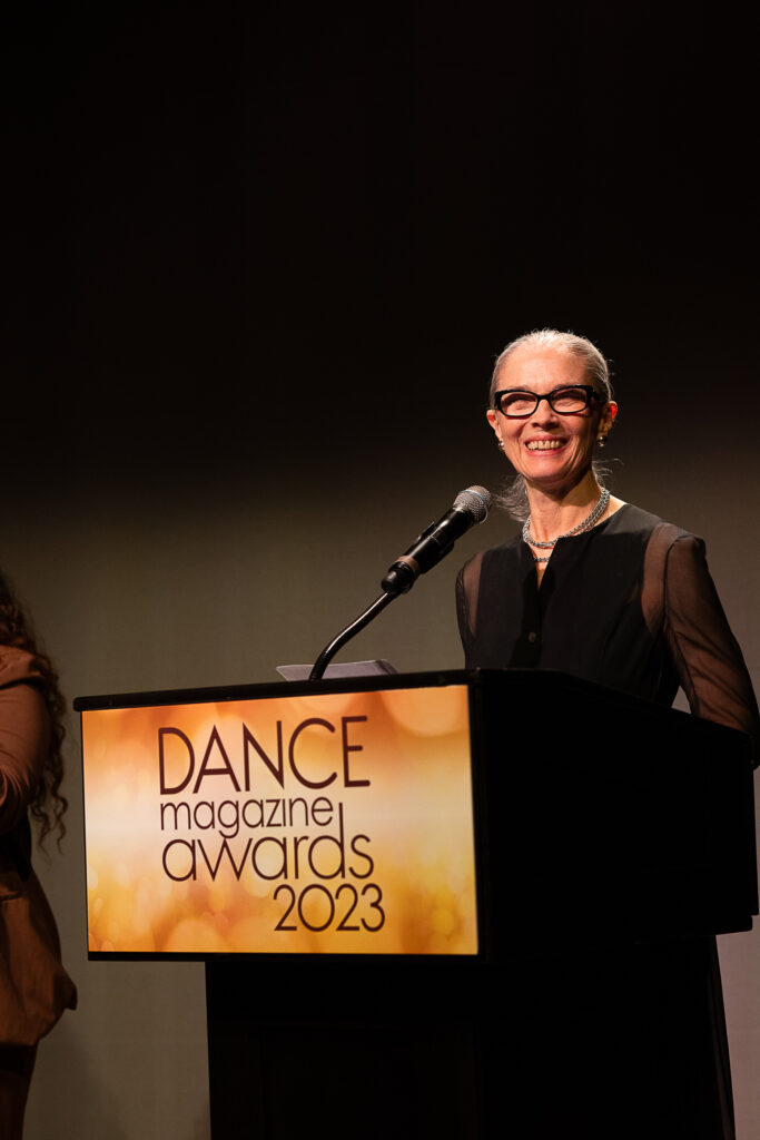 Janet Eilber, an elegant white woman with her silver hair pulled neatly back, smiles warmly behind a podium with a sign that reads "Dance Magazine Awards 2023."