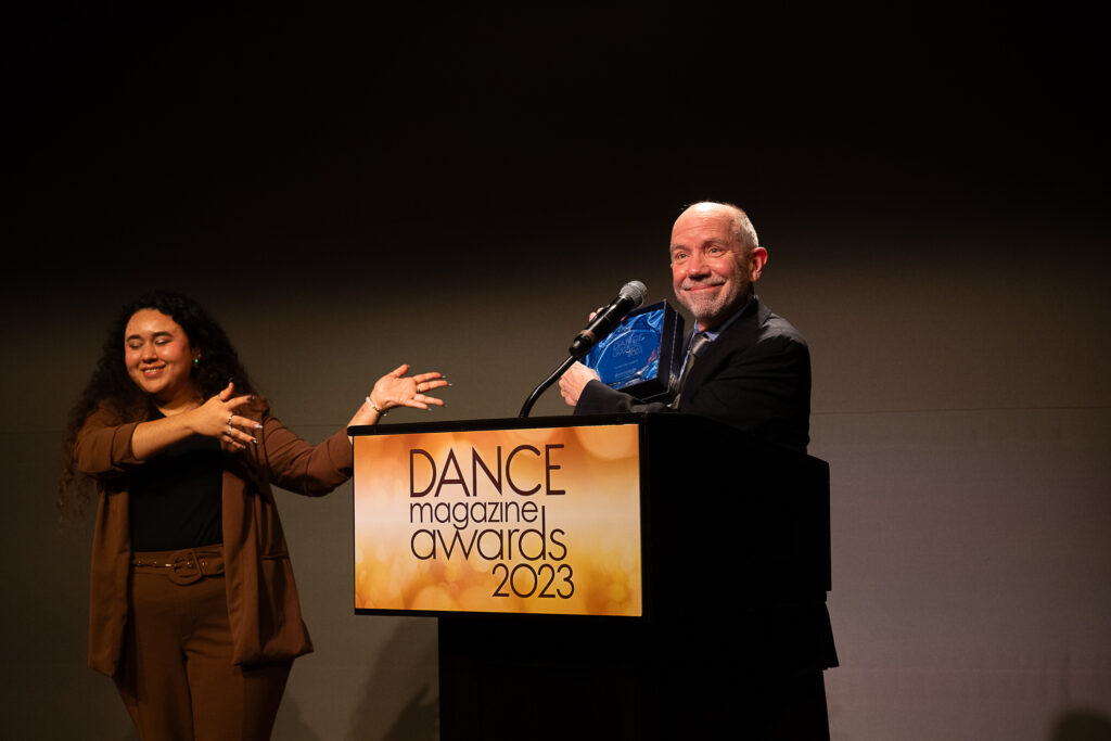 Norton Owen, an older white man, gives a closed mouth smile as he proudly holds up the box containing his glass Dance Magazine Award. The ASL interpreter to his right smiles as she gestures toward him. He stands behind a podium with a sign that reads "Dance Magazine Awards 2023."