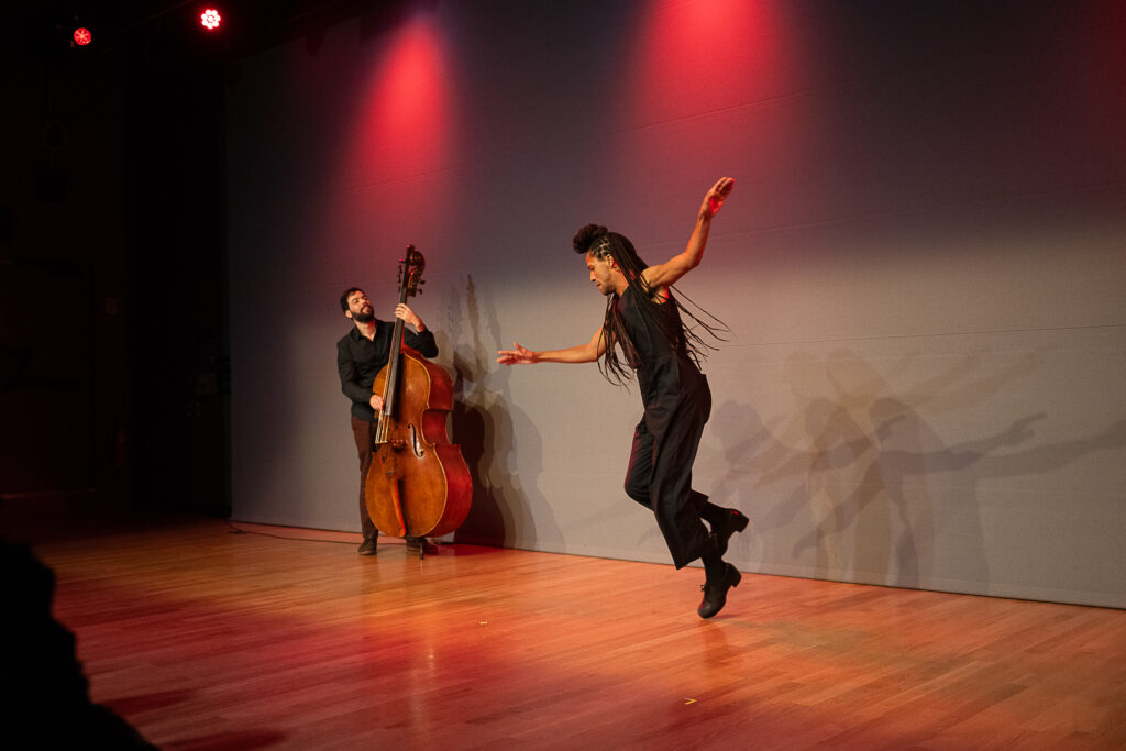 Leonardo Sandoval, a Brazilian man with long braids piled atop his head and flowing behind him, hovers just above the floor as he does a pullback in his tap shoes. With his gracefully extended arms, he seems about to take flight. Upstage, a man strums a standing bass, watching Sandoval.