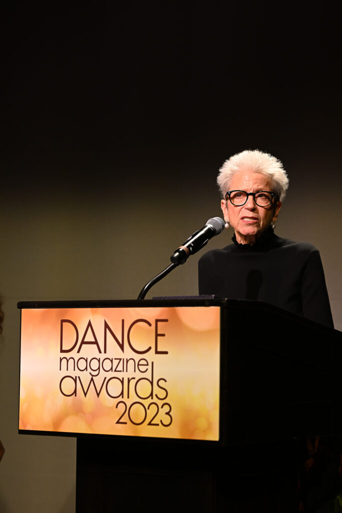 Jody Gottfried Arnhold, an older white woman with a shock of short, white hair and black rimmed glasses, speaks intently into a microphone. She is at a podium with a sign that reads, "Dance Magazine Awards 2023."
