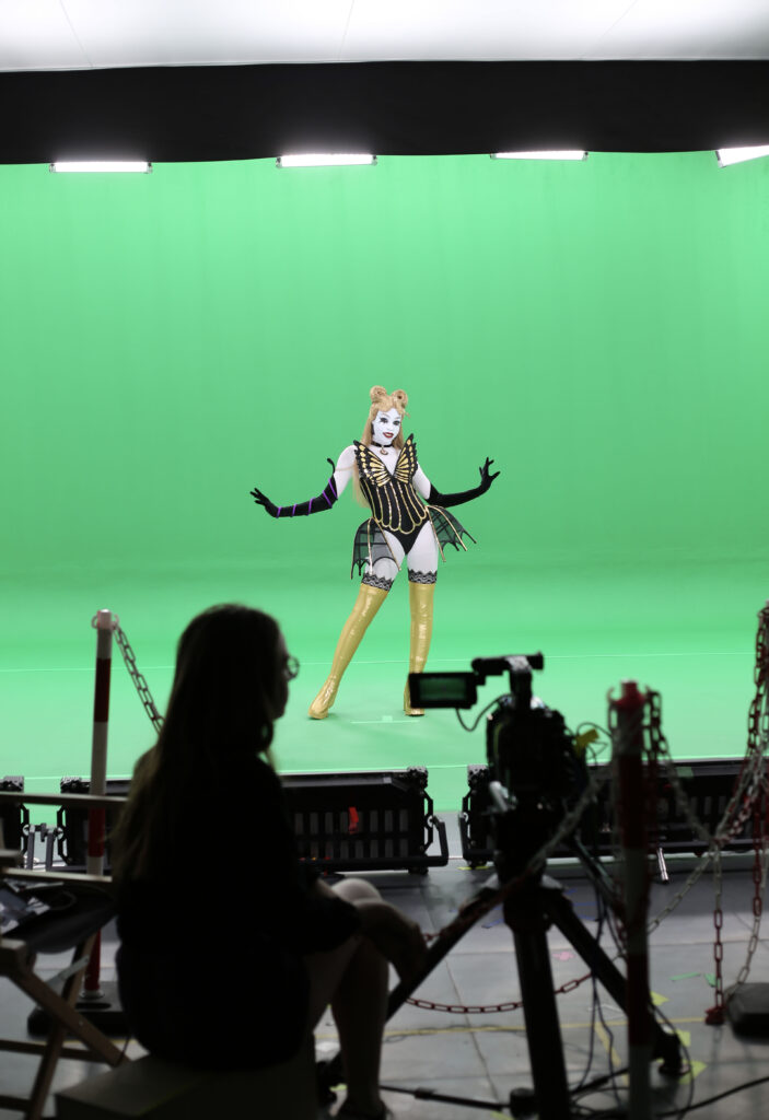 A dancer in a highly stylized butterfly costume, bright wig, and airbrushed makeup dances against a green screen. In the foreground, the outlines of cameras and someone in a director's chair are visible in silhouette.