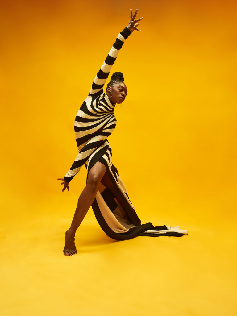 a woman wearing a bold black and white striped dress posing in front of a bright yellow background 