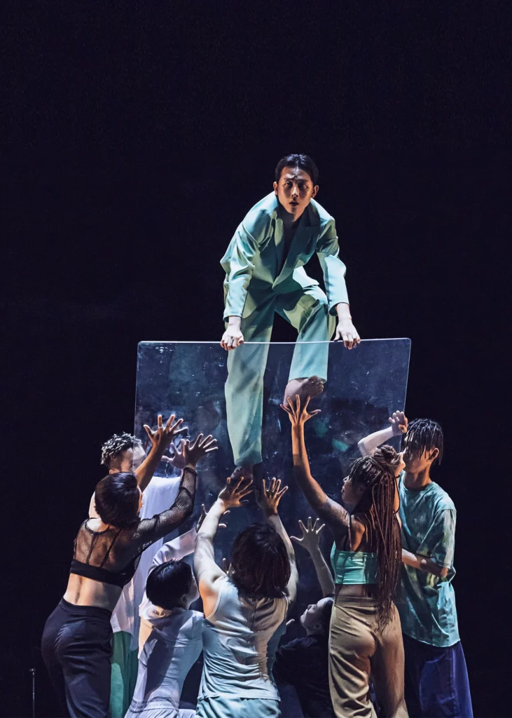 A dancer climbs to the top of a see-through panel as it teeters forward, supported by a clump of seven dancers on either side of the panel.
