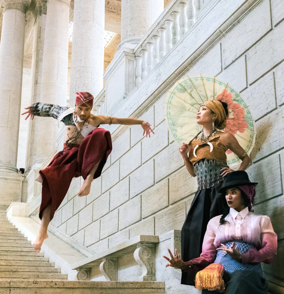 Three dancers are photographed on a staircase lined with white stone and pillars. They are dressed in contemporized versions of old-fashioned Filipino costumes. One leans against a wall under a parasol, while another is captured midair, arms spread wide. The third is seat, hands splayed as though casting a spell.