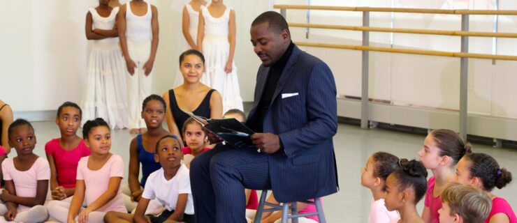 Robert Battle sits on a chair reading aloud from a book. Young students in dance clothes sit arrayed around him, listening.