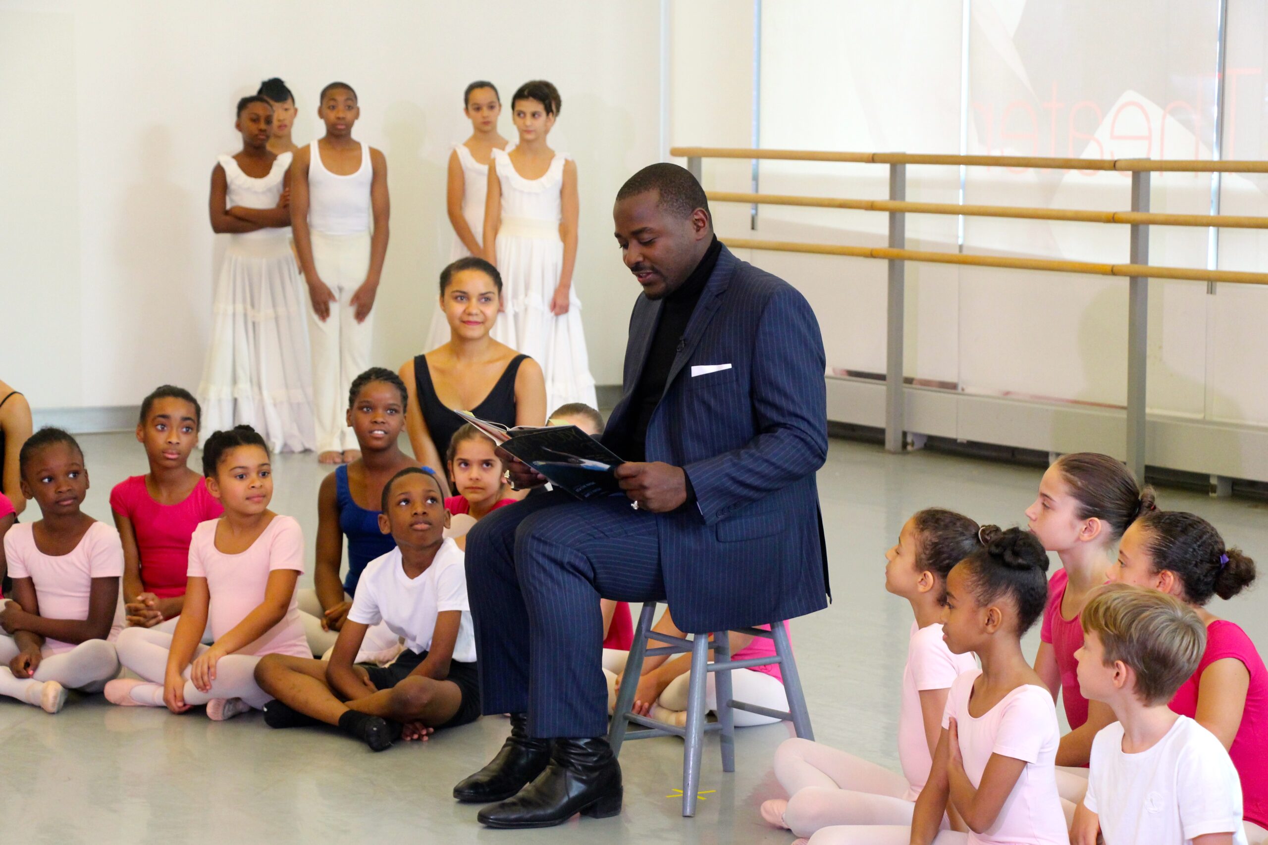 Robert Battle sits on a chair reading aloud from a book. Young students in dance clothes sit arrayed around him, listening.
