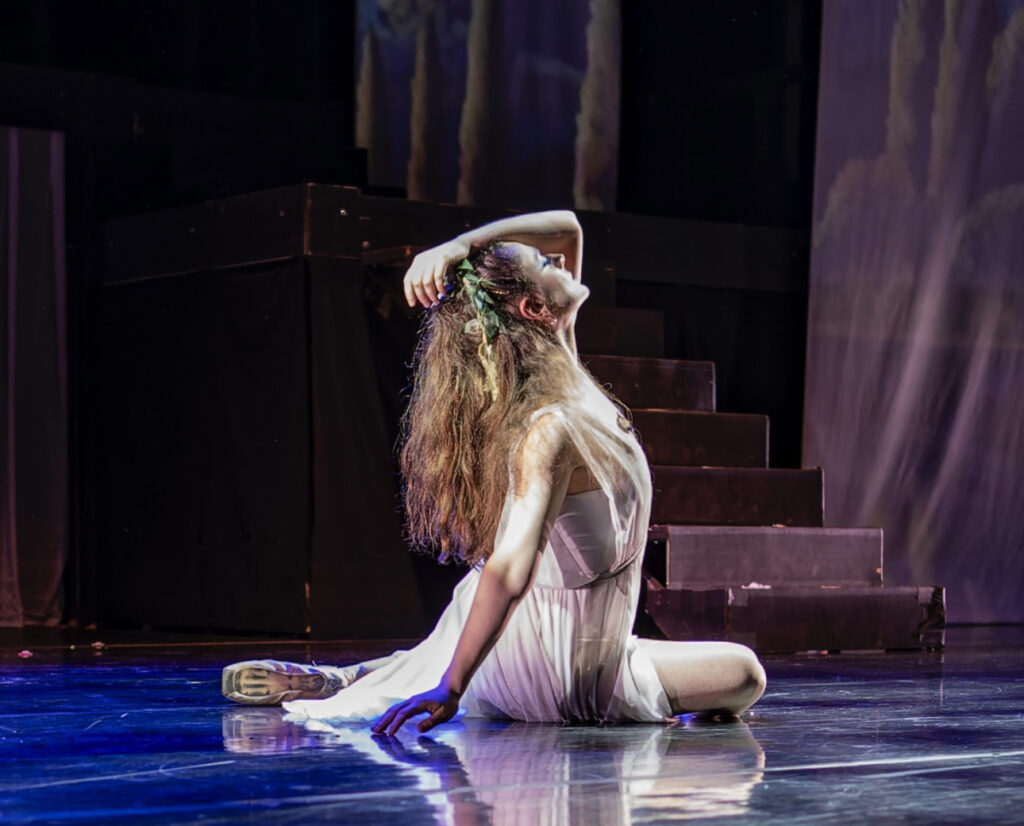 a dancer on stage sitting on the floor with one above her head. she had her hair down and is wearing a flowy white dress