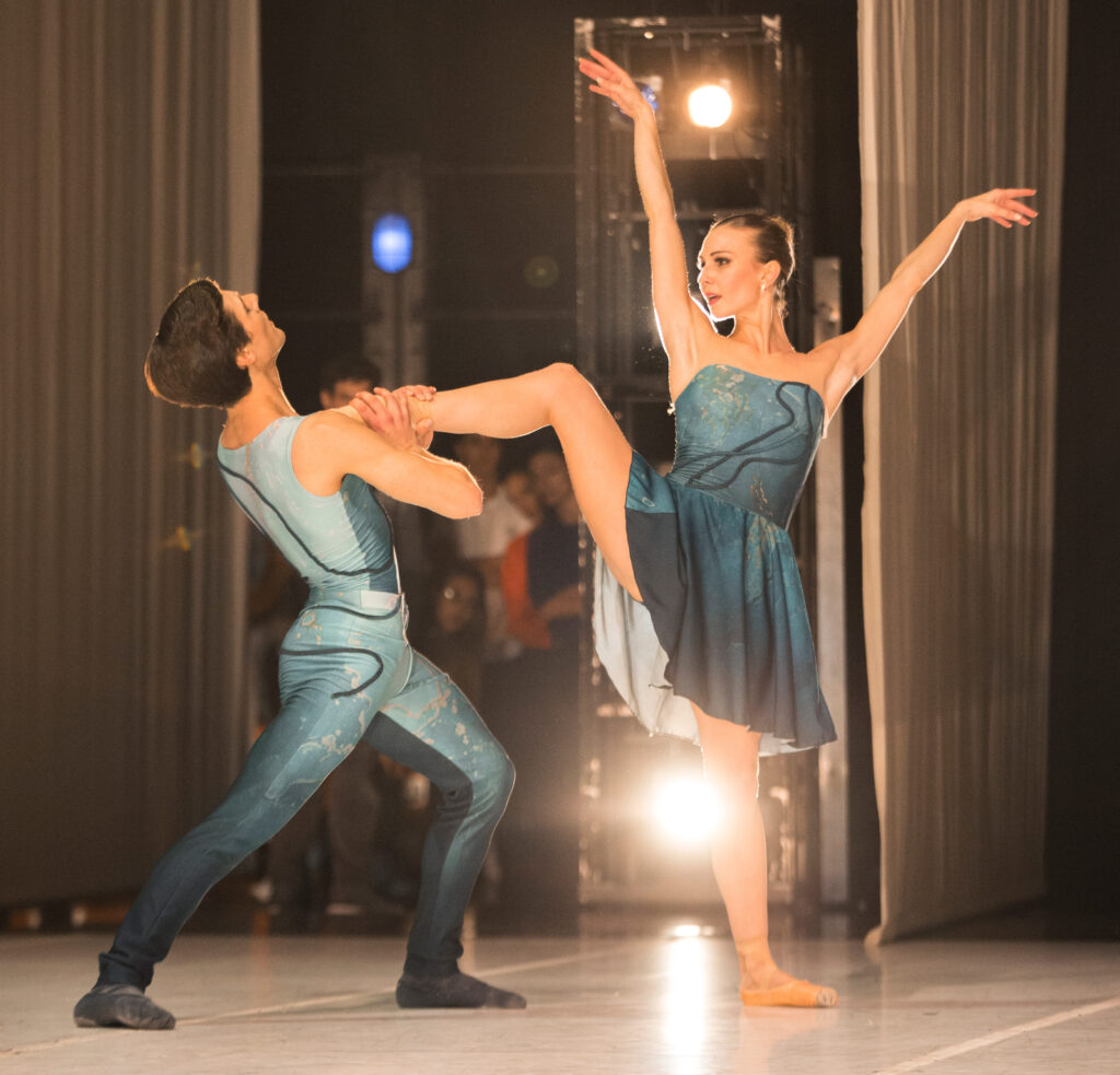 The view of a pas de deux from the wings. A woman in pointe shoes raises one leg in side attitude, arms in a V by her head. A male dancer lunges toward her while leaning back, hands cradling her extended foot to his chest. Both wear turquoise, the man in a fitted unitard, the woman in a skirt that flows to just above her knees.