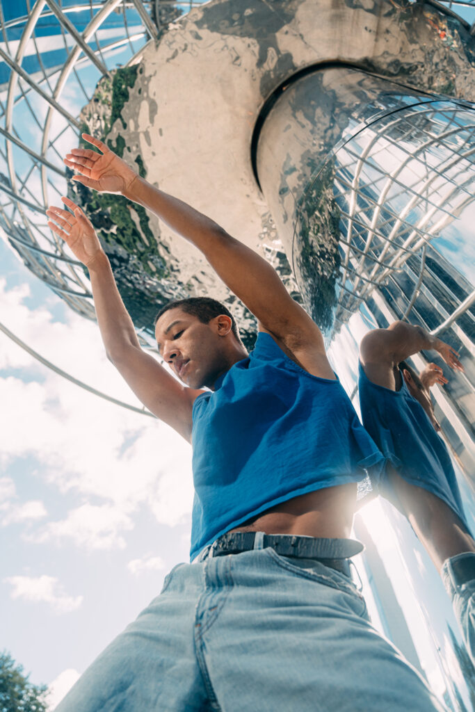 a male dancer wearing jeans and a tank top posing with his arms over his head against a mirrored background 