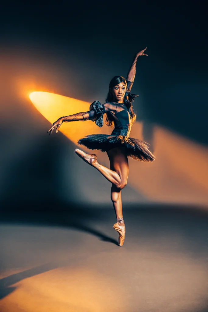 A Black ballerina poses en pointe against a dramatically lit grey backdrop. She is in parallel, knees squeezed together as she lifts one foot behind her. She looks over her shoulder to the camera, arms in an elegant "L' shape. She wears a black tutu with dramatic poufs at the upper arms and pointe shoes that match her skin color.